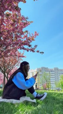 Diş teli takan kadın telefonda selfie çekiyor arka planda sakura açıyor.