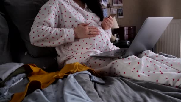 Glücklich Schwangere Frau Sieht Niedliche Babykleidung Die Bett Liegt Stock-Filmmaterial