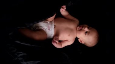Siyah arka planda yatan bir bebek, kısmen siyah bir bezle kaplı, tek koluyla yukarı bakıyor..