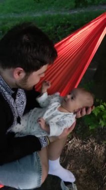 Hamakta oturan bir adam, arka planda ağaçlar olan bir yerde bebeği tutuyor..