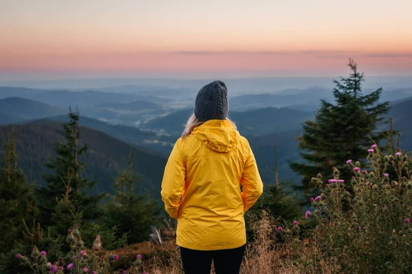 stock image Woman wearing yellow jacket and knit hat and looking at mountain range during sunset. Hiking in mountains