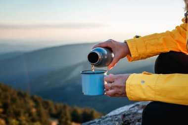 Kadın turist termostan dağlardaki seyahat bardağına sıcak su dolduruyor. Yürüyüş sırasında yiyecek içecek.