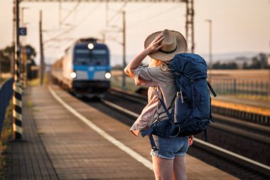 Trenle seyahat et. Tren istasyonunda duran ve treni bekleyen şapkalı bir kadın.