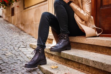 Trençkot giyen ve ayak bileği çizmeli bir kadın şehirde merdivenlerde oturuyor. Modaya uygun siyah deri ayakkabı. Sokak modası