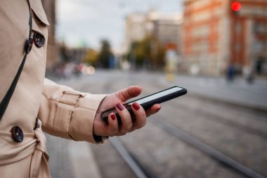 Şehir caddesinde otobüs durağında bekleyen bir kadın akıllı telefonunu kontrol ediyor. Kadın elinde cep telefonu.