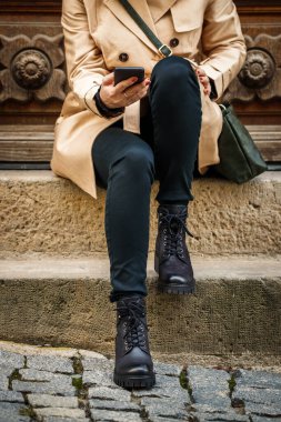 Merdivenlerde oturan ve şehirde akıllı telefon kullanan bir kadın. Trençkot giyen şık bir kadın, siyah kot pantolon ve deri botlar. Sokak tarzı