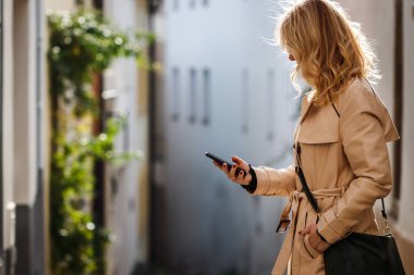 Şehir caddesinde akıllı telefonunu kullanan trençkotlu bir kadın. Cep telefonundaki kadın mesajları