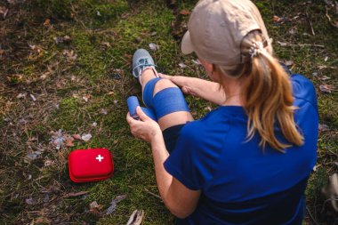 First aid after hiking accident. Injured hiker putting elastic bandage to her knee. Tendon problems or sprain knee during trekking in nature clipart