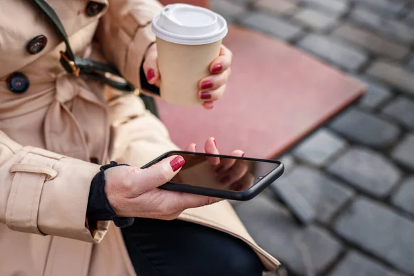 Stock image Businesswoman resting on bench and using smart phone during city break on street. Woman text messaging on mobile phone and drinking coffee