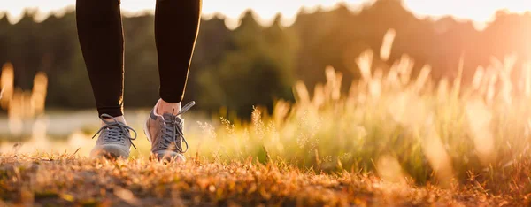 Spor ayakkabılı ve tozlu bir kadın gün batımında doğada koşuyor. Koşan kadın bacakları ve çimenlerdeki güneş ışığı panoramik açıdan görülüyor. Spor ve aktif yaşam tarzı kavramı