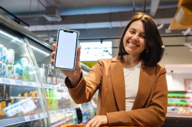 Elinde alışveriş arabası olan gülümseyen bir kadın markette beyaz ekranlı bir telefon tutuyor.