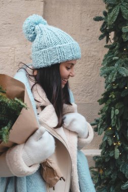 Kışın gülümseyen güzel bir kadın elinde Noel ağacı buketiyle dışarıda yürüyor.