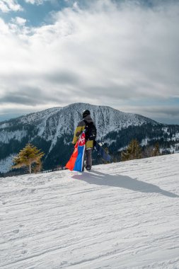 Slovakya bayrağı taşıyan snowboardcu kayak merkezinin yamacında arka planda güzel dağlar var.