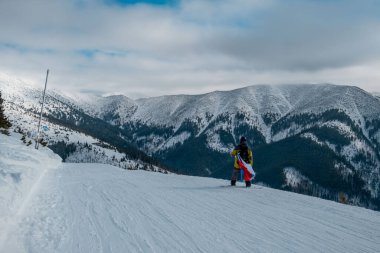 Slovakya bayrağı taşıyan snowboardcu kayak merkezinin yamacında arka planda güzel dağlar var.