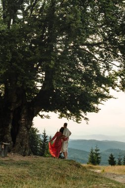 Karpatlar 'ın dağlarını ve günbatımını gören büyük bir kayın ağacının altında duran bir çift.
