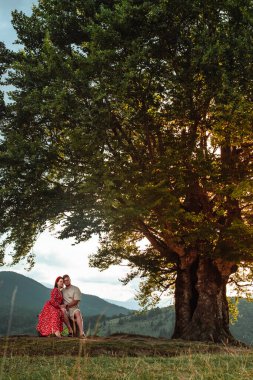 a couple sits on a bench by a big old beech tree with a view of the carpathians mountains