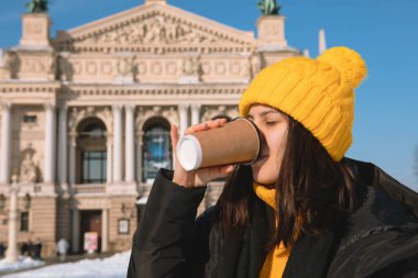 Elinde tek kullanımlık fincanla arka planda Lviv opera binası içen genç bir kadın.