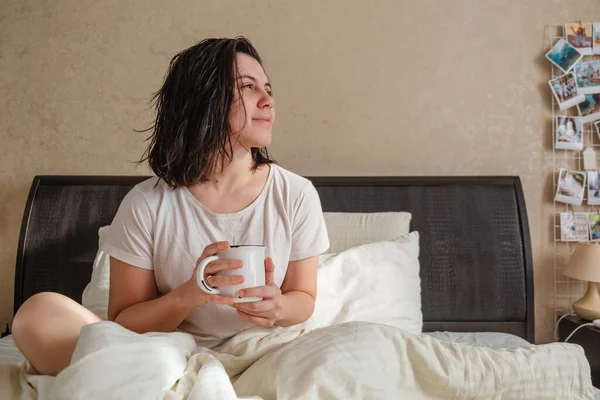 A woman with wet hair is sitting on a bed holding a cup of hot drink