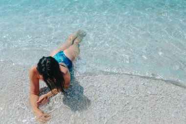 Mavi mayo giymiş güzel bir kadın kumsalda güneşlenirken deniz suyuna uzanmış.