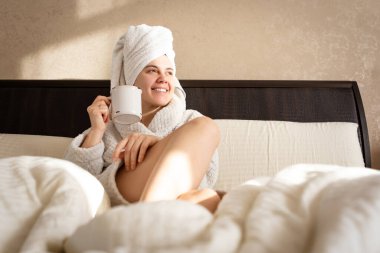 In a white dressing gown with a towel on her head, a woman holds a cup on a bed