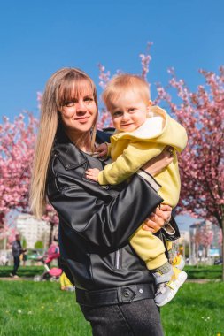 Gülümseyen anne, baharda pembe kiraz çiçeklerinin yanında oğlunu elleriyle tutuyor.
