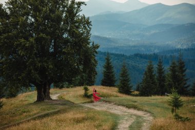 woman in red dress on the path near large old beech tree with lush green leaves in Carpathian mountains in summer time. Landscape