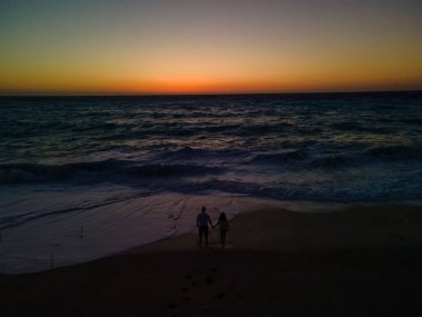 Deniz kıyısında durup gün batımında güçlü dalgalara bakan çiftlerin silueti.