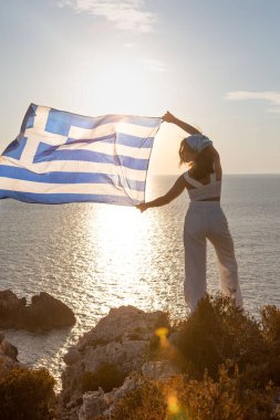 Yunan bayrağı taşıyan kadın arka planda Lefkada adasının deniz fenerinde gün batımına bakıyor.