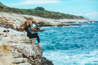 Genç, sıska bir kadın uçurumun kenarında oturmuş denize bakıyor. Boşluğu kopyala