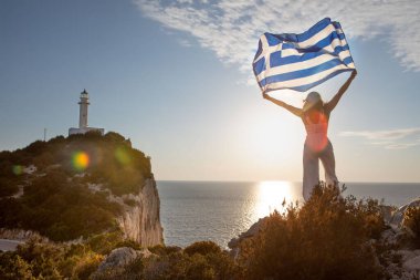 Yunan bayrağı taşıyan kadın arka planda Lefkada adasının deniz fenerinde gün batımına bakıyor.