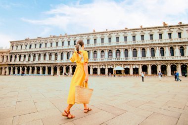 Venedik Saint Marco Meydanı 'nda saman çantalı zarif sarı elbiseli bir kadın yürüyor. Yaz tatili. Boşluğu kopyala