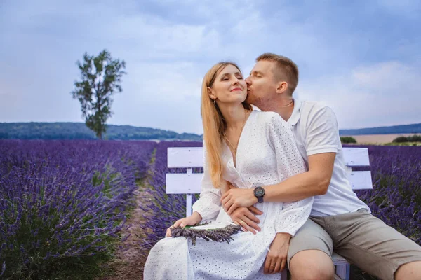 In the center of a lavender field, a couple in love sits on a bench and shares a kiss