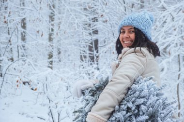 Karlı bir ormanda, kışın ortasında, bir kadın elinde karlı bir Noel ağacı, mutlu bir şekilde gülümsüyor.