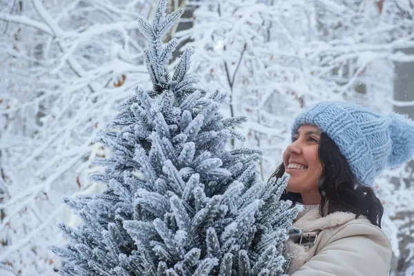 Gülümseyen kadın ellerindeki karlı Noel ağacına bakar.