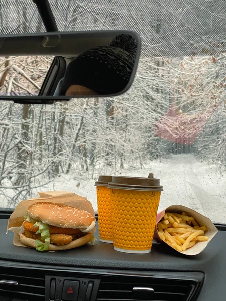 hamburger, french fries and 2 coffees on dashboard car. road through the winter forest