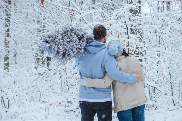 couple hugs in snowy forest. man has Christmas tree on his shoulder. back view