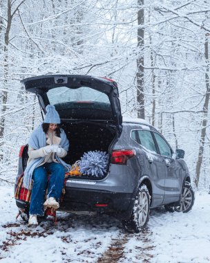 Kış ormanının ortasında, genç bir kadın Noel ağacıyla arabanın bagajında oturuyor.