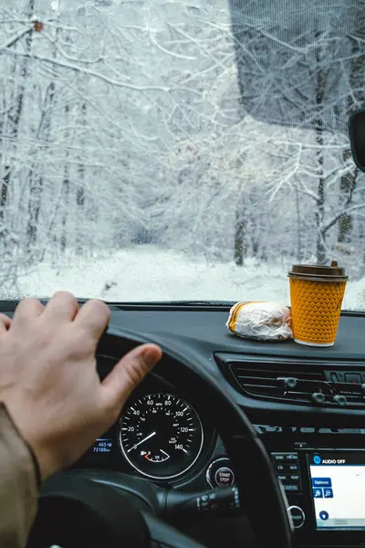mans hand on steering wheel of car, hamburger and coffee on dashboard