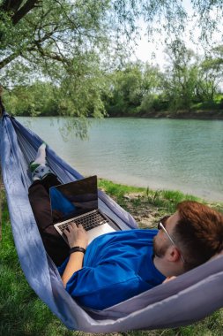 Genç bir adam nehir kıyısındaki bir hamakta yatarken dizüstü bilgisayarıyla çalışıyor.