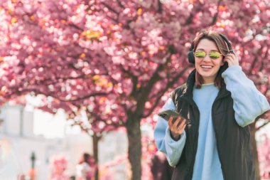 Kadın müzik aşığı telefonundaki şarkıları kontrol ediyor. Arka planda sakura açıyor.