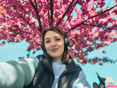 Güzel, gülümseyen bir kadın kulaklıkla müzik dinliyor. Bahar zamanı çiçek açan sakura ağacının altında selfie çekiyor.