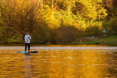 A person paddleboarding on a calm lake surrounded by autumn foliage. clipart