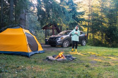 Huzurlu bir ormanda kamp yaparken arka planda Çadır SUV arabasının yanındaki kamp ateşine bakıyor.