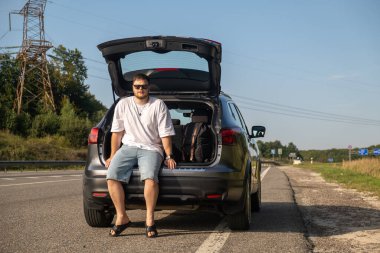 Genç adam güneşli bir yolda arabasının yanında dinleniyor.