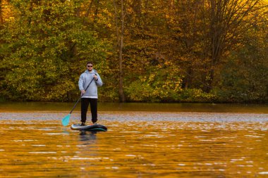 A person paddleboarding on a calm lake surrounded by autumn foliage. clipart