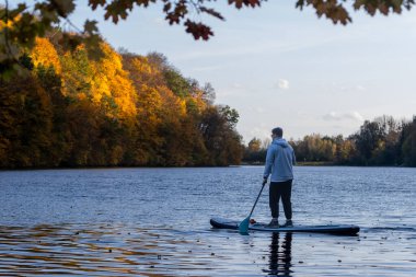 A person paddleboarding on a calm lake surrounded by autumn foliage. clipart