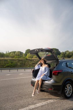 Beyaz elbiseli ve şapkalı bir kadın arabanın bagajında oturuyor, güneşli bir günün tadını çıkarıyor.