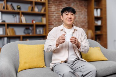 Bir adam, parlak sarı yastıkları olan yumuşak gri bir kanepeye rahatça oturur, kitap raflarıyla dolu zarif bir oturma odasında çayını yudumlar ve parlak bir ambiyansı vardır..