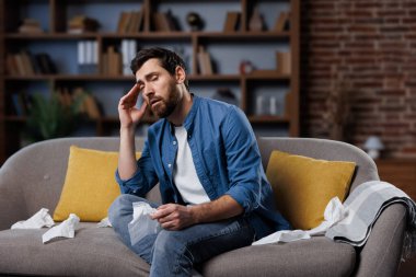 Hasta, hasta, kötü hisseden, baş ağrısı çeken bir adam şık bir oturma odasında rahat bir koltukta otururken kendini hasta hissediyor gibi görünüyor. Alnını ovalarken bir elinde mendil tutuyor.