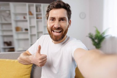 Selfie çekiyor, video çekiyor, sakallı neşeli bir adam mutluluğunu ifade ediyor. Parlak bir oturma odasında selfie çekerken başparmağını kaldırıyor. Sade uzayda bitkiler ve sarı yastıklar var..